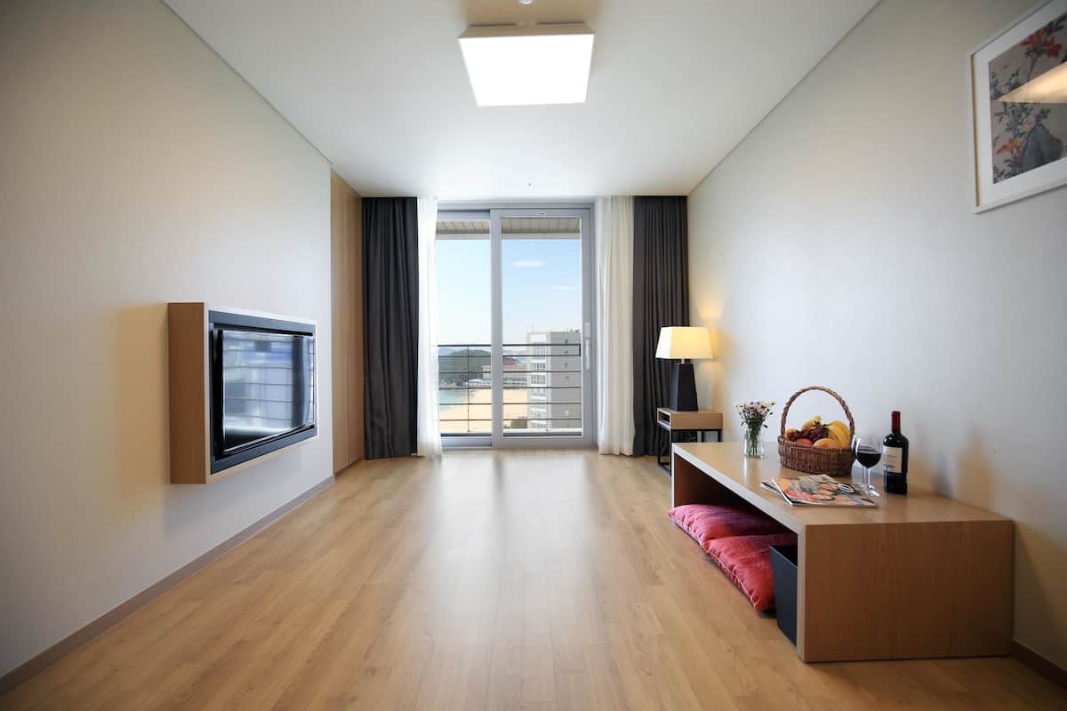 A modern hotel room with a large window, a flat-screen TV, a desk with a lamp, and a small table with a basket of fruit and a bottle of wine.