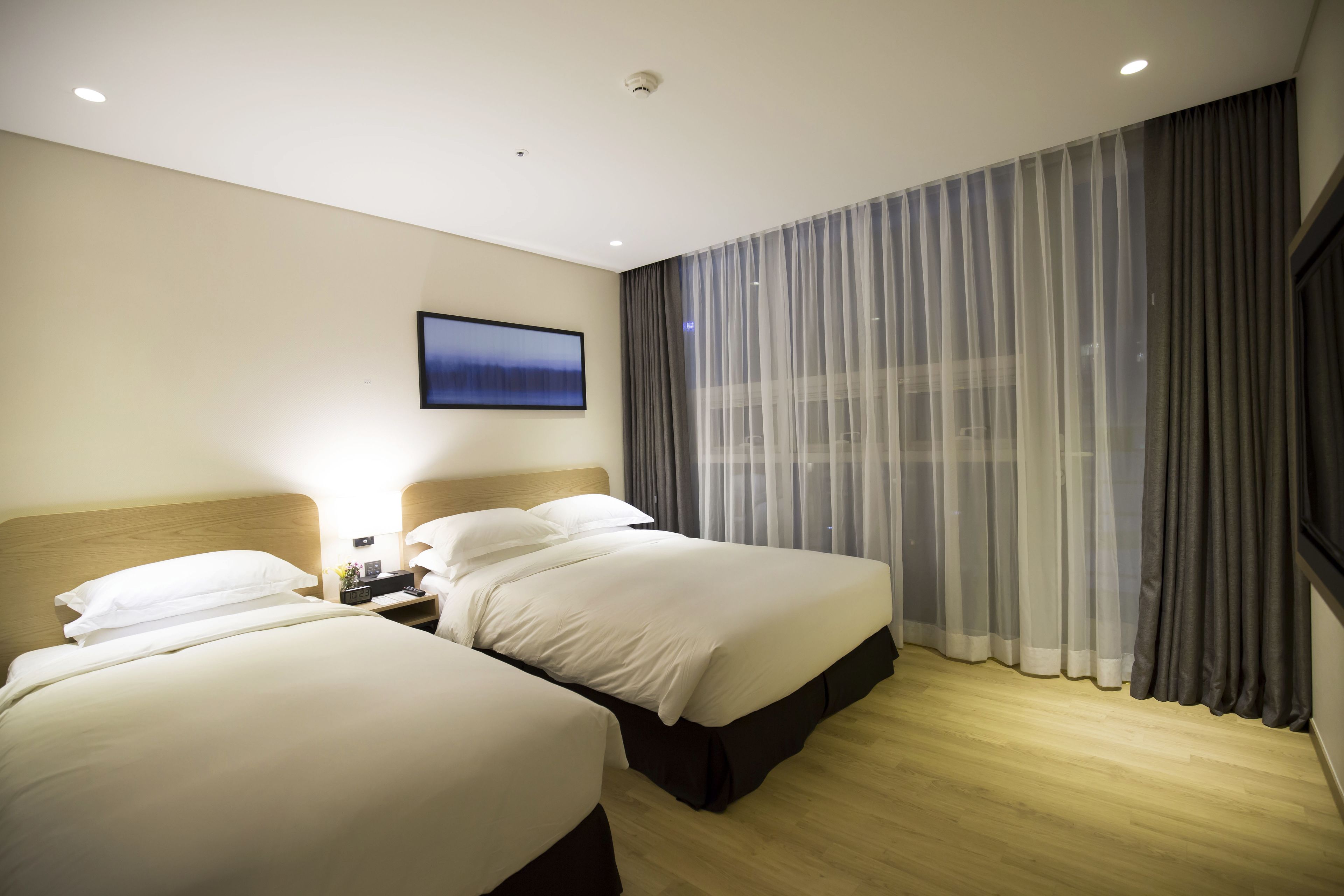 Une chambre d’hôtel avec deux lits, un téléviseur à écran plat fixé au mur et une grande fenêtre avec des rideaux légers.