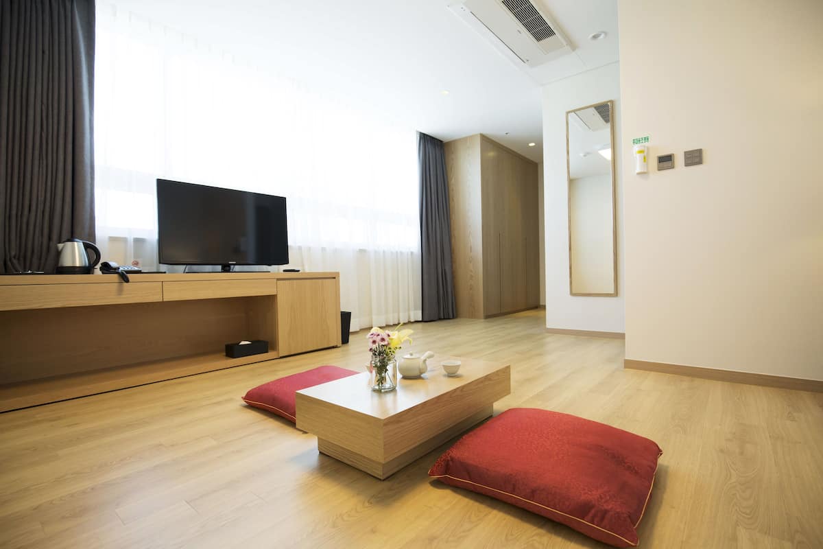 A modern hotel room with a wooden floor, a flat-screen TV, a coffee table with a vase of flowers, and red cushions.