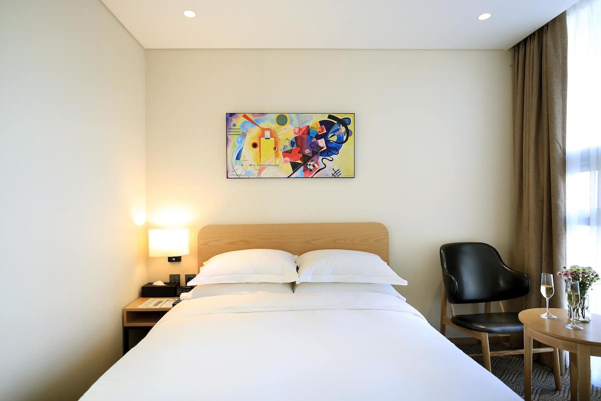 A hotel room with a bed, a chair, a table with a glass of wine, and a painting on the wall.