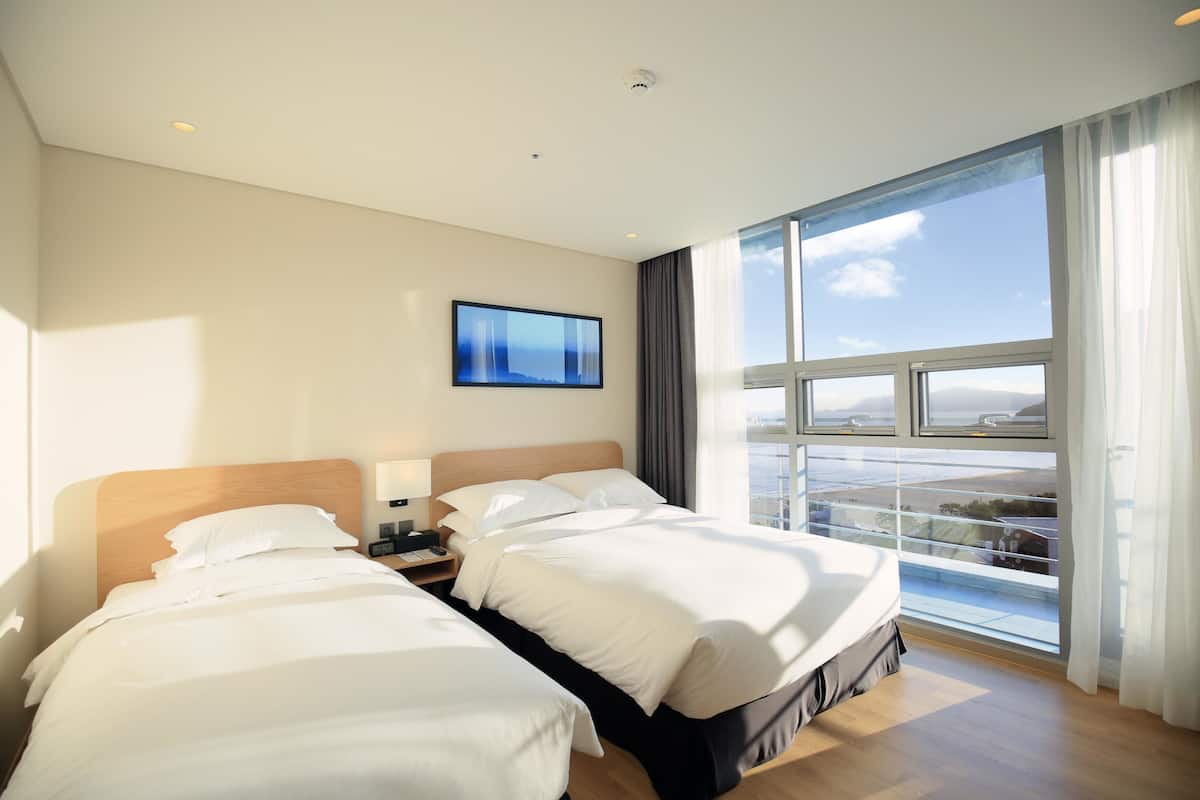 A hotel room with two beds, a large window with a view, and a flat-screen TV on the wall.