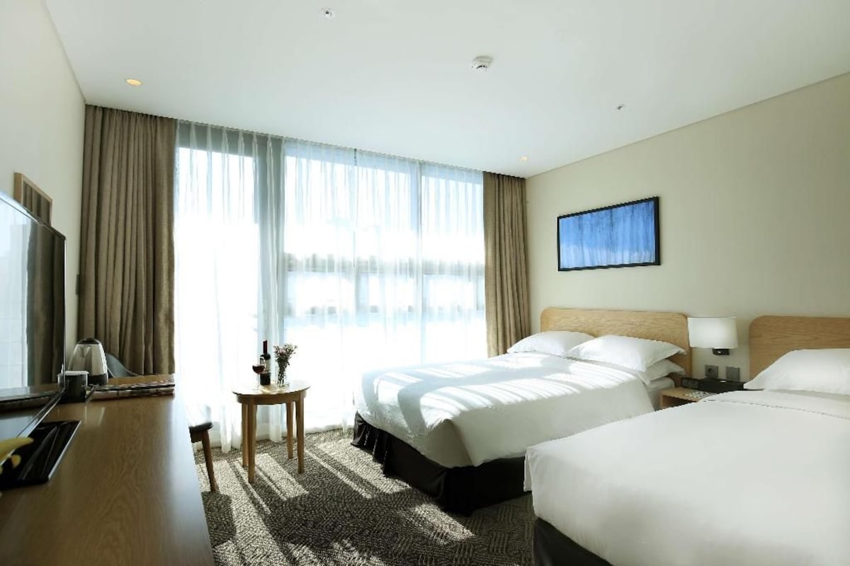A hotel room with a large bed, a bedside table, a lamp, a TV, and a window with curtains.