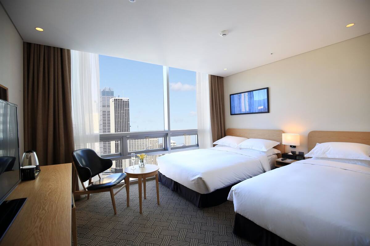 A hotel room with two beds, a desk with a chair, a small table with a vase, and a large window offering a city view.