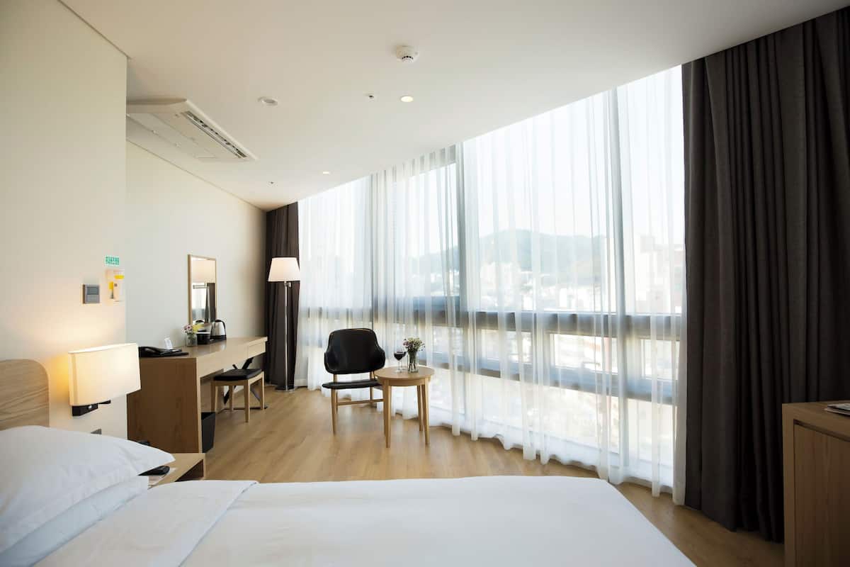 A hotel room with a large bed, a desk with a chair, a small table with a lamp, and a view of the city through sheer curtains.
