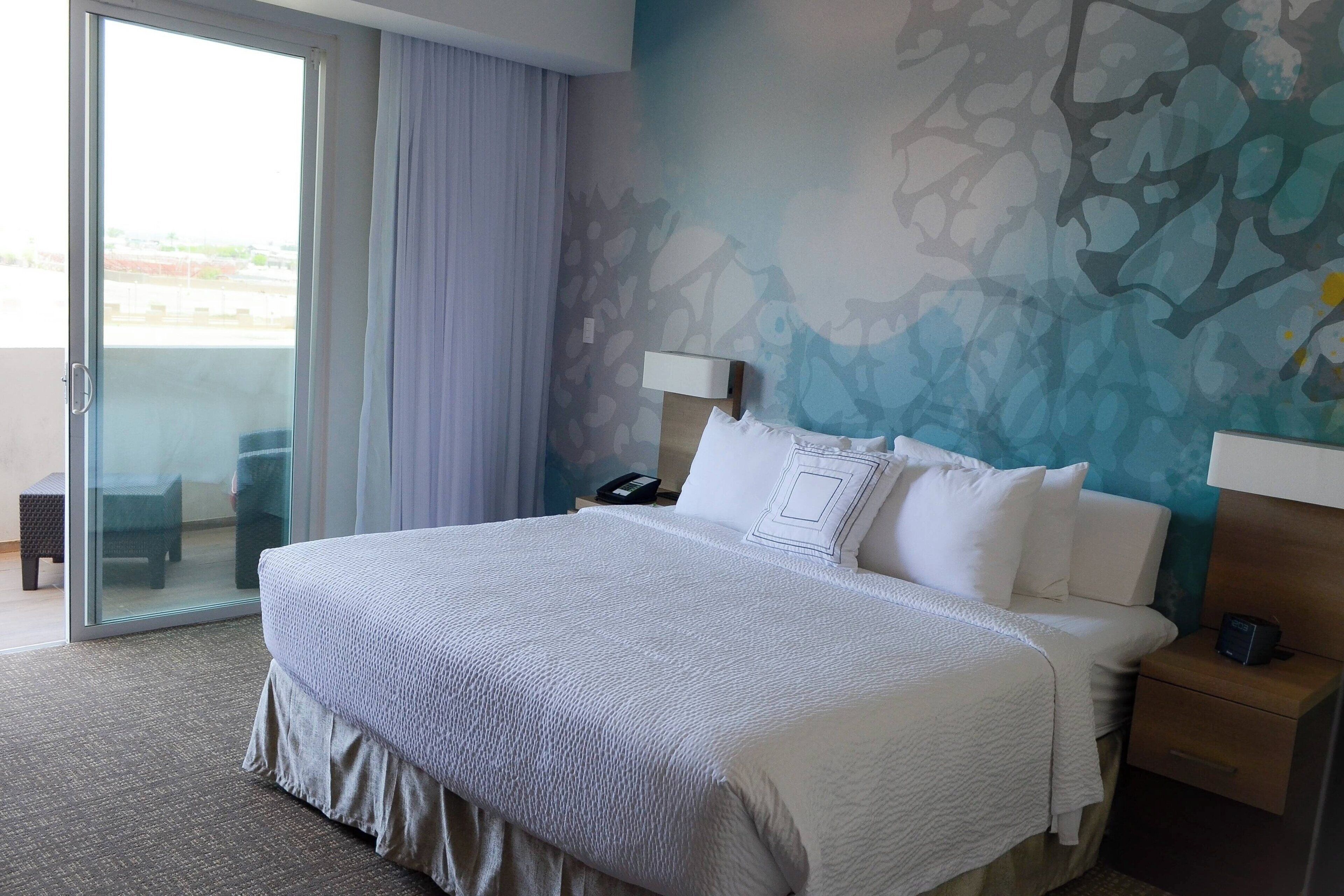 Ein Hotelzimmer mit einem großen Bett, einem Nachttisch, einer Lampe, einem Telefon und Blick durch eine Glastür.