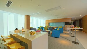 A modern hotel lobby with a central bar area, seating arrangements, and a large window providing natural light.