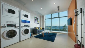 A laundry room with stacked washing machines, a gray sofa with yellow pillows, and a large window offering a city view.