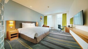 A hotel room with a large bed, bedside tables, a desk, a chair, a flat-screen TV, and a large window with curtains.