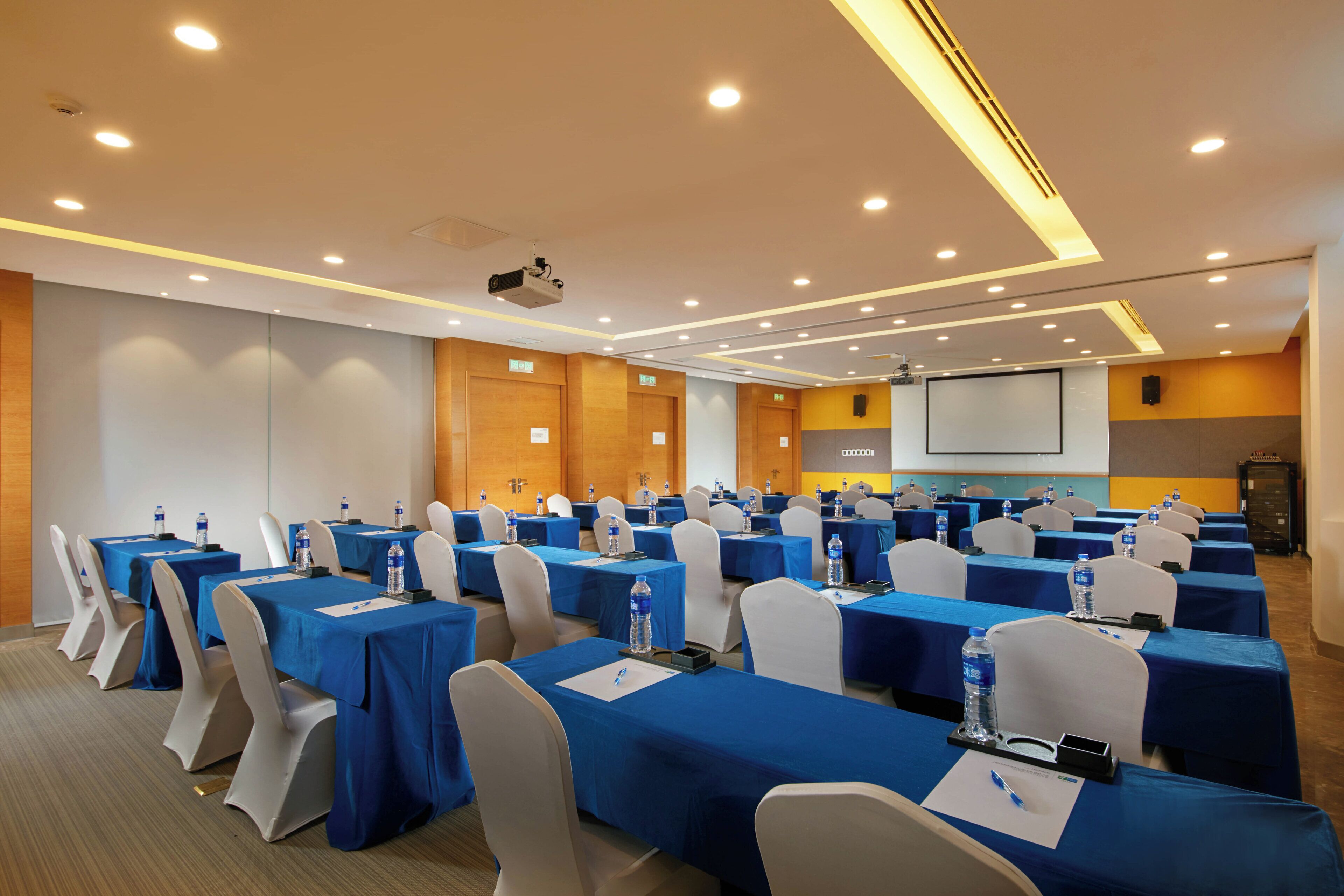 Un salón de conferencias con varias mesas, sillas y una pantalla para proyector.