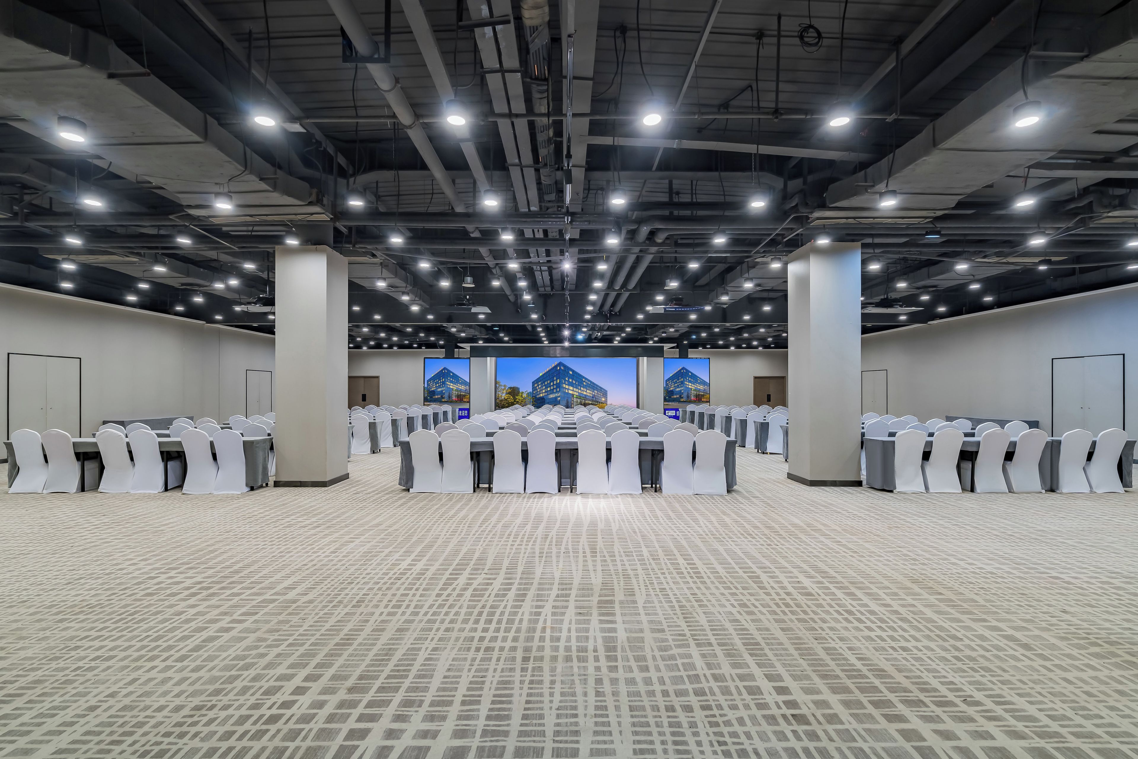 Une grande salle de conférence avec de nombreuses tables et chaises disposées en rangées, un plafond haut orné de luminaires et une vue sur les bâtiments à travers de grandes fenêtres.