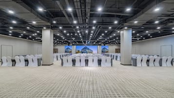 Une grande salle de conférence avec de nombreuses tables et chaises disposées en rangées, un plafond haut orné de luminaires et une vue sur les bâtiments à travers de grandes fenêtres.
