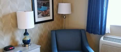 Quarto de hotel com uma cama, uma mesinha de cabeceira com abajur, um quadro de Spokane, uma poltrona azul e uma janela com cortinas.