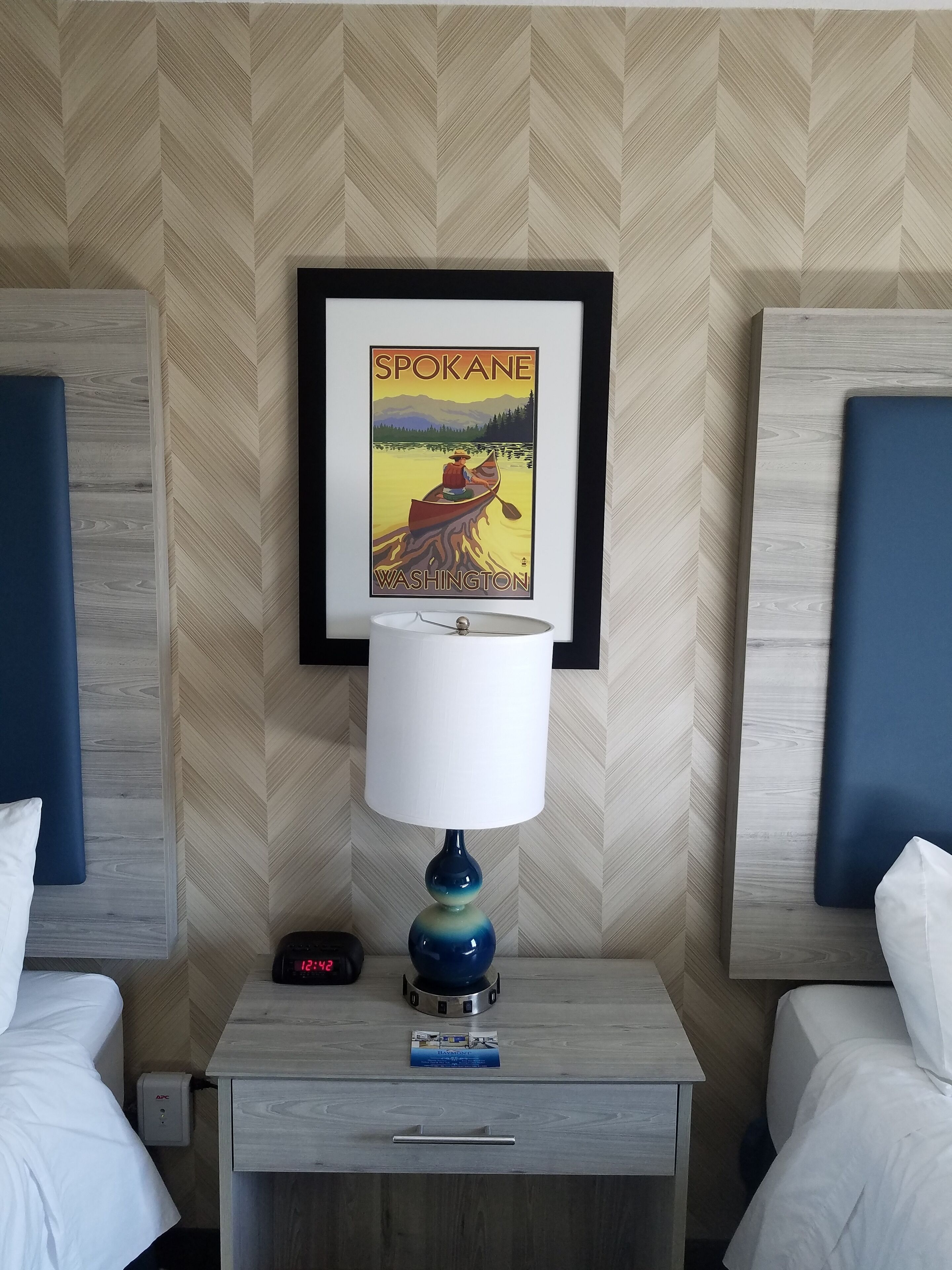 A hotel room with a bed, a nightstand with a lamp, a framed poster of Spokane, Washington, and a digital clock displaying 12:42.