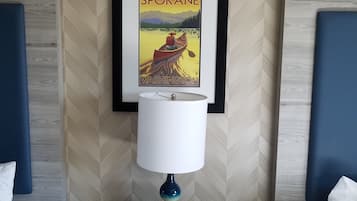 Een hotelkamer met een bed, een nachtkastje met een lamp, een lijst met een foto van Spokane, Washington, en een digitale klok waarop 12:42 staat.