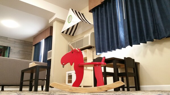 A room with a dining table, chairs, a rocking horse, a flag, and a window with curtains.