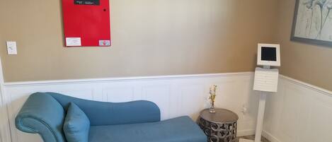 A room with a blue sofa, a red fire alarm, a small round table with a vase, and a white wall-mounted device.