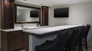 Een moderne keuken met een ingebouwd spoelbak, een grote spiegel en een flatscreen televisie boven de werkruimte. Er staan vier barkrukken met metalen stiksels.