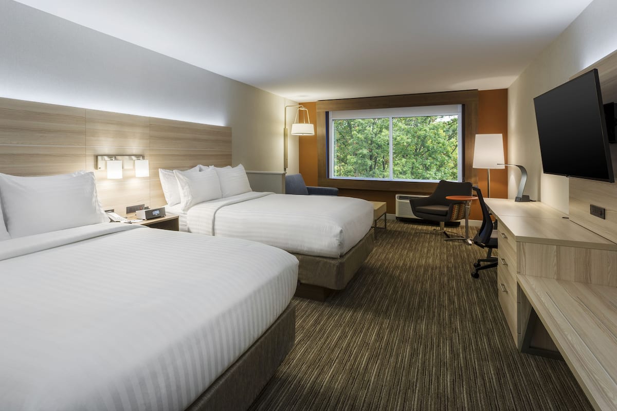 Quarto de hotel com duas camas, uma escrivaninha, uma cadeira, uma televisão e uma janela com vista para árvores.