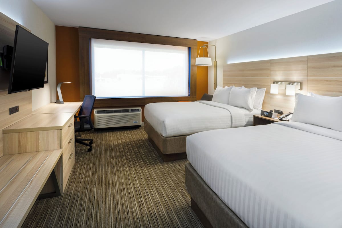 Quarto de hotel com duas camas, uma escrivaninha, uma cadeira, uma televisão, um aquecedor e uma janela com persianas.