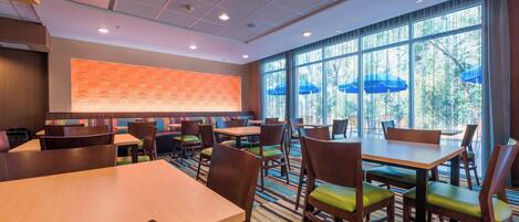 A hotel dining area with multiple tables and chairs, large windows with blue umbrellas, and a patterned wall.