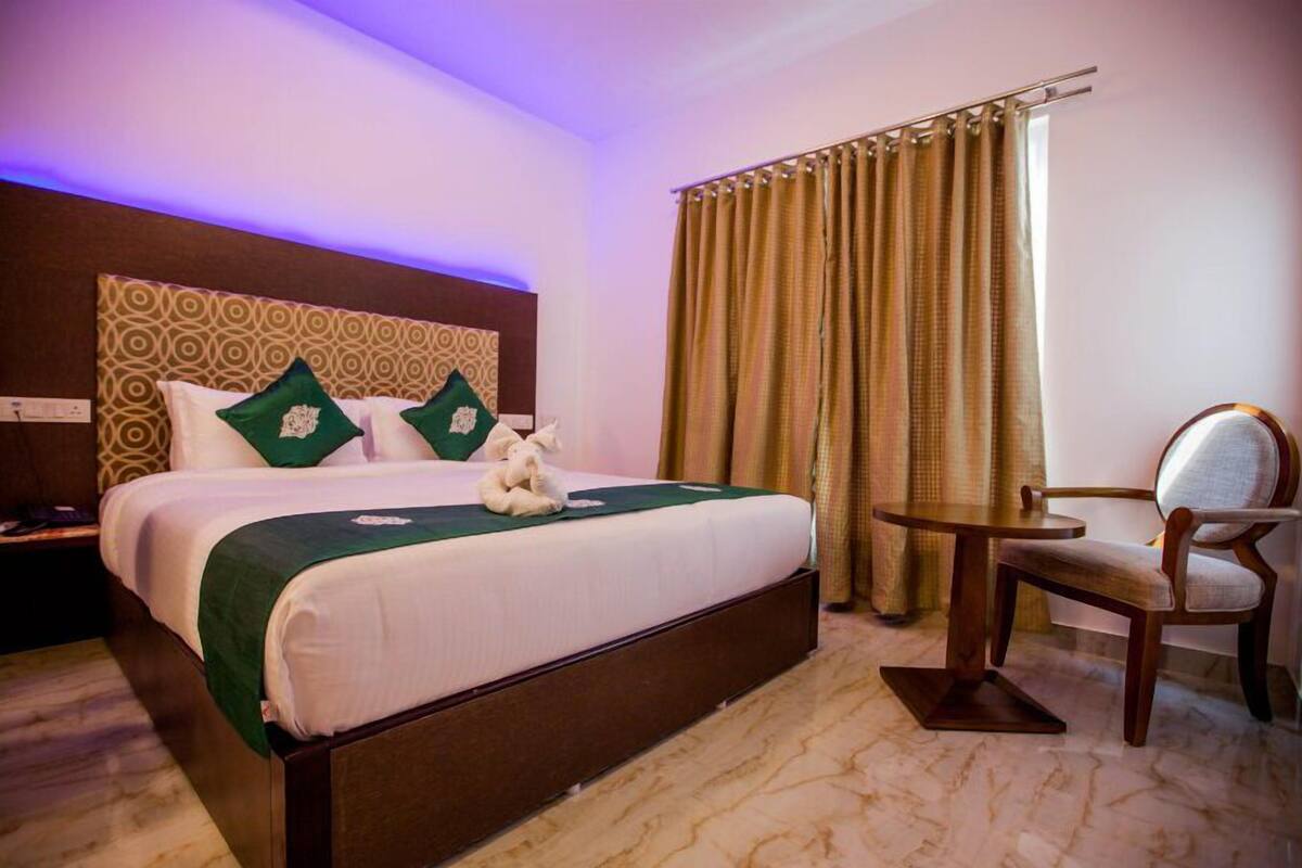 A hotel room with a large bed, a wooden headboard, a green throw, two pillows, a teddy bear, a round table, a chair, and a curtain.