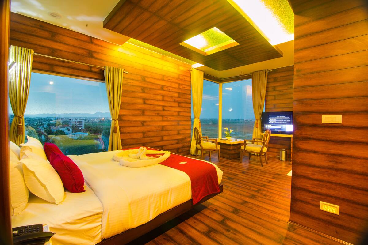 A hotel room with a large bed, wooden walls, a view of the city, and a desk with a chair.