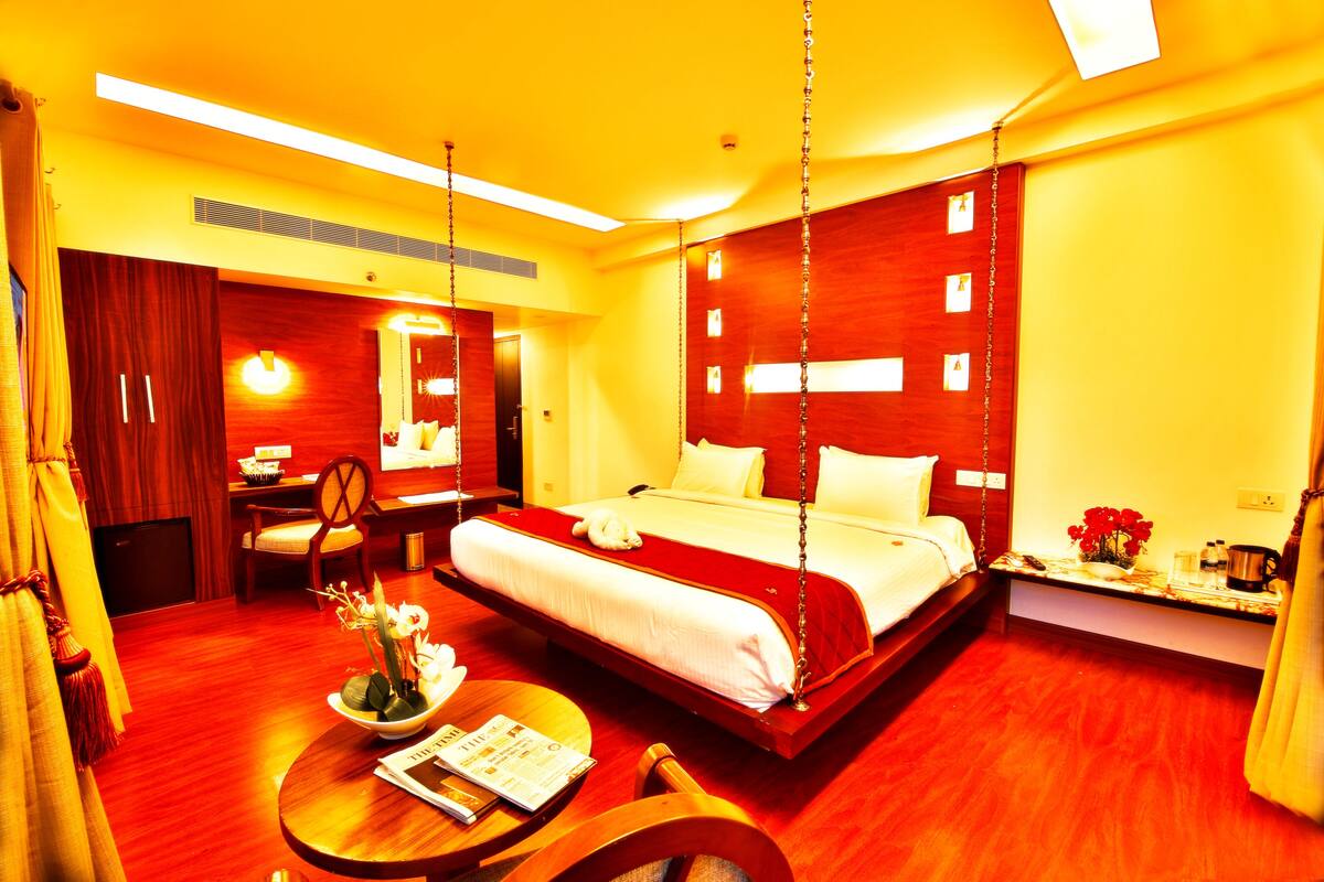 A hotel room with a large bed, a desk, a chair, a small table with a vase of flowers, and a bedside table with a lamp and a clock.