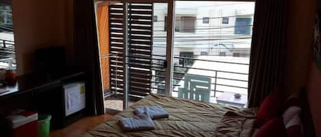 A hotel room with a bed, a desk with a computer, a green trash bin, and a window with blinds.