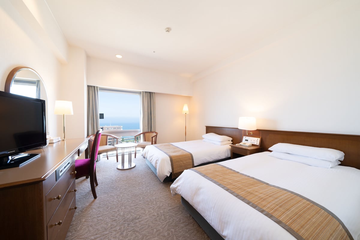 A hotel room with two beds, a desk with a TV, a chair, and a view of the ocean.