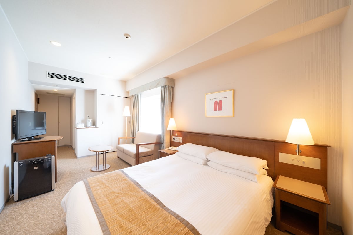A hotel room with a large bed, bedside tables with lamps, a television, a mini-fridge, and a seating area with a chair and table.