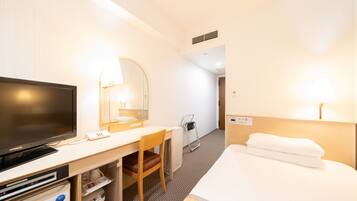 A hotel room with a bed, a desk with a TV, a chair, a desk lamp, and a bedside table.