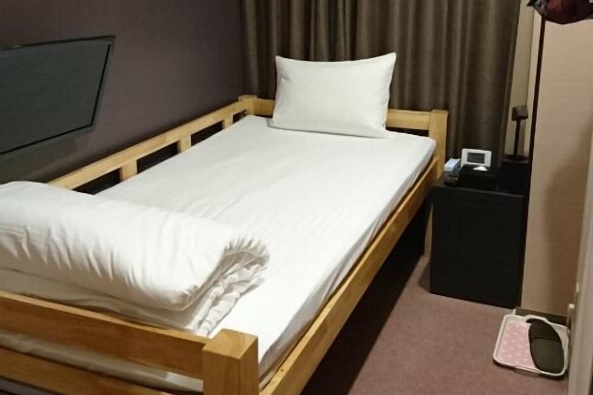 A single bed with a wooden frame, a white mattress, and a pillow. There is a small black nightstand with a lamp and a radio beside the bed. The room has a carpeted floor and a dark curtain in the background.
