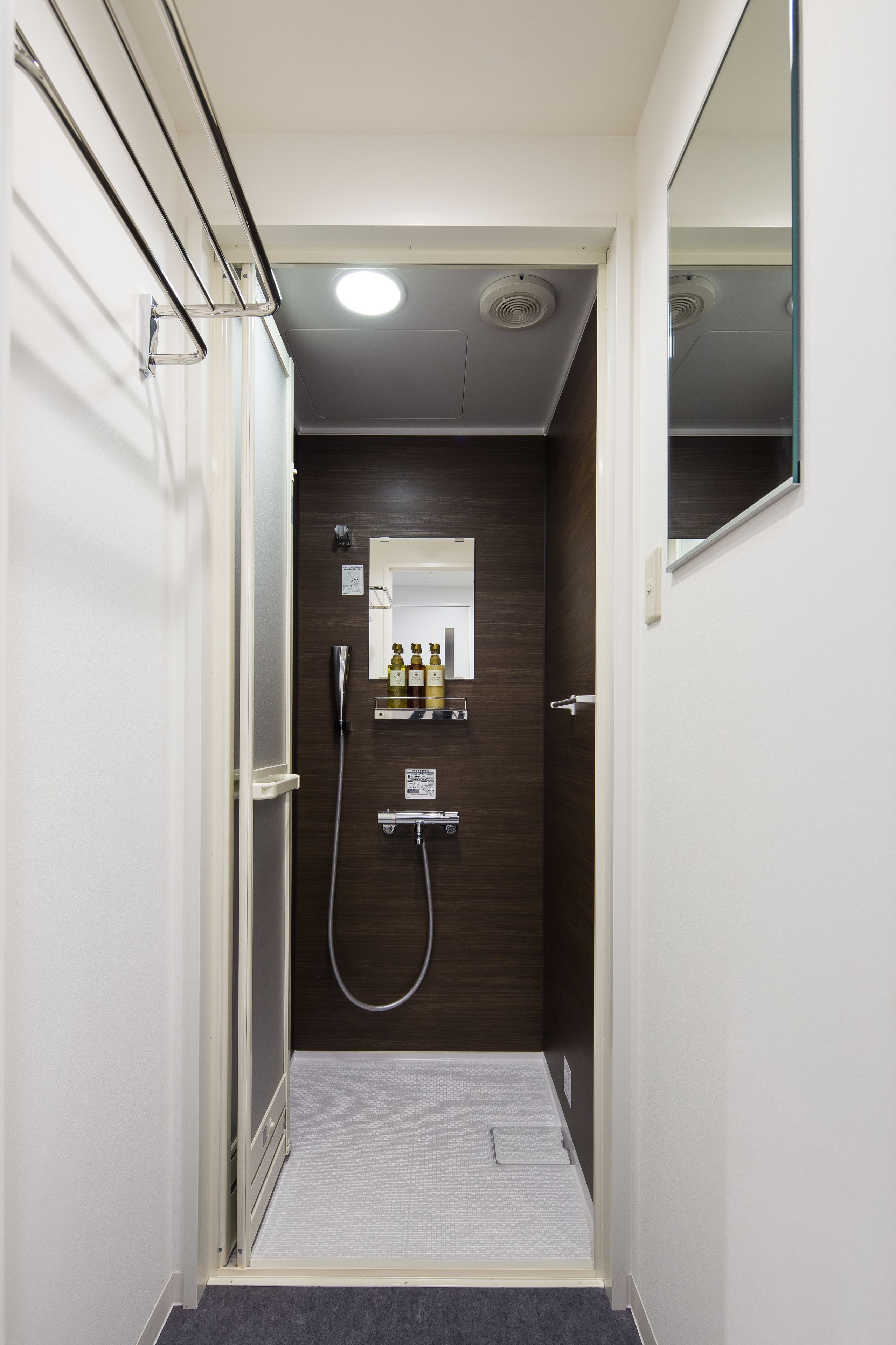 A narrow, modern bathroom with a shower, a mirror, and a shelf with toiletries.