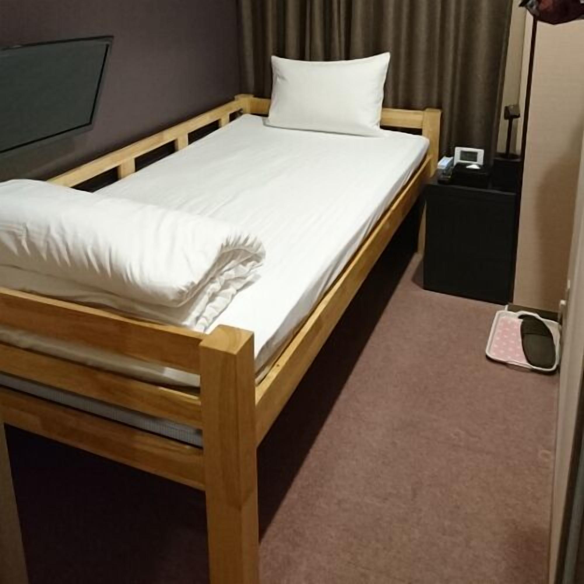 A single bed with a wooden frame, a white mattress, and a pillow. There is a small black nightstand with a lamp and a phone. The room has a carpeted floor and a dark curtain.