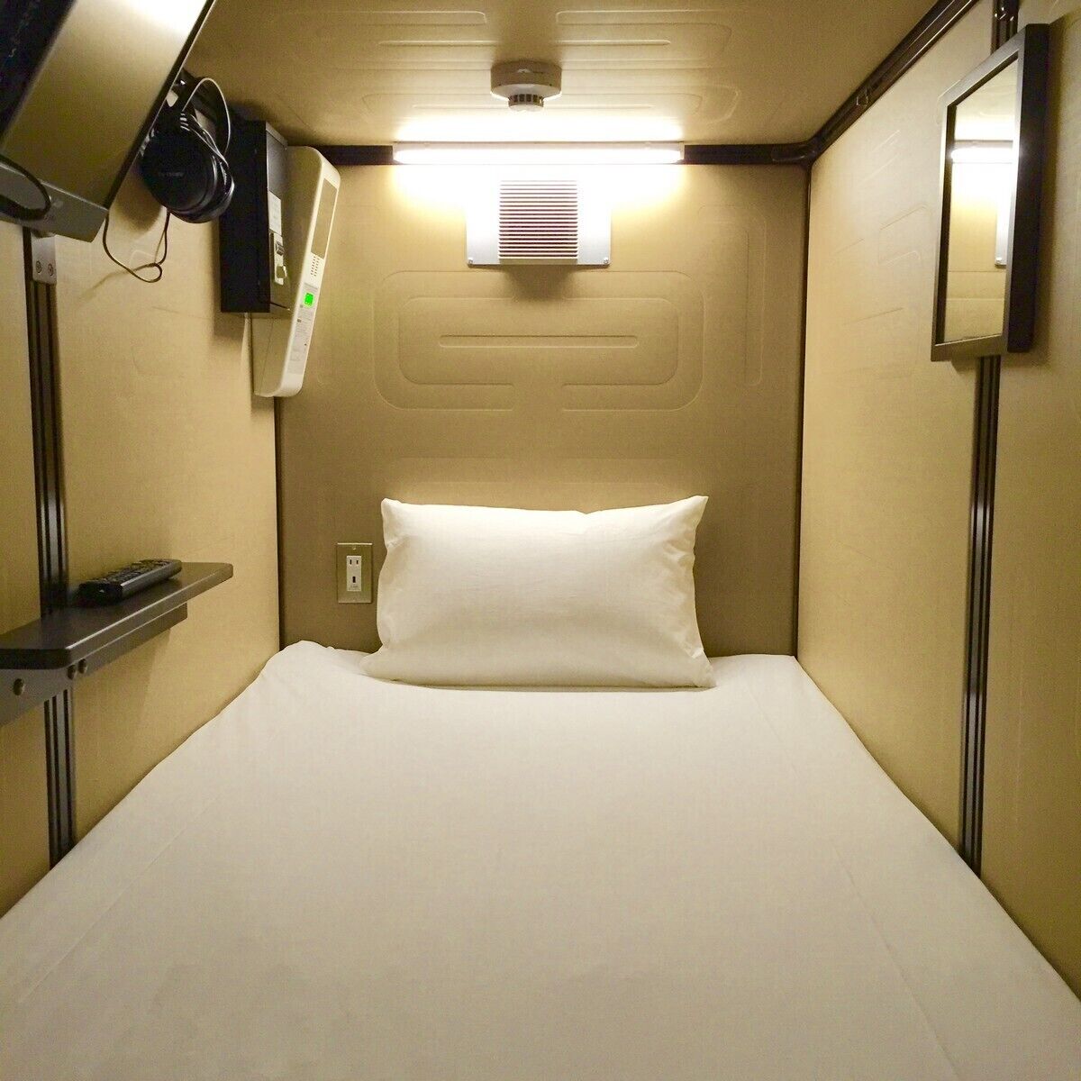 A compact hotel room with a bed, a bedside table, a wall-mounted heater, and a mirror.