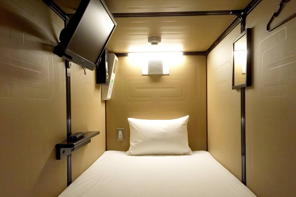 A narrow hotel room with a single bed, a mounted television, and a wall-mounted air conditioning unit.