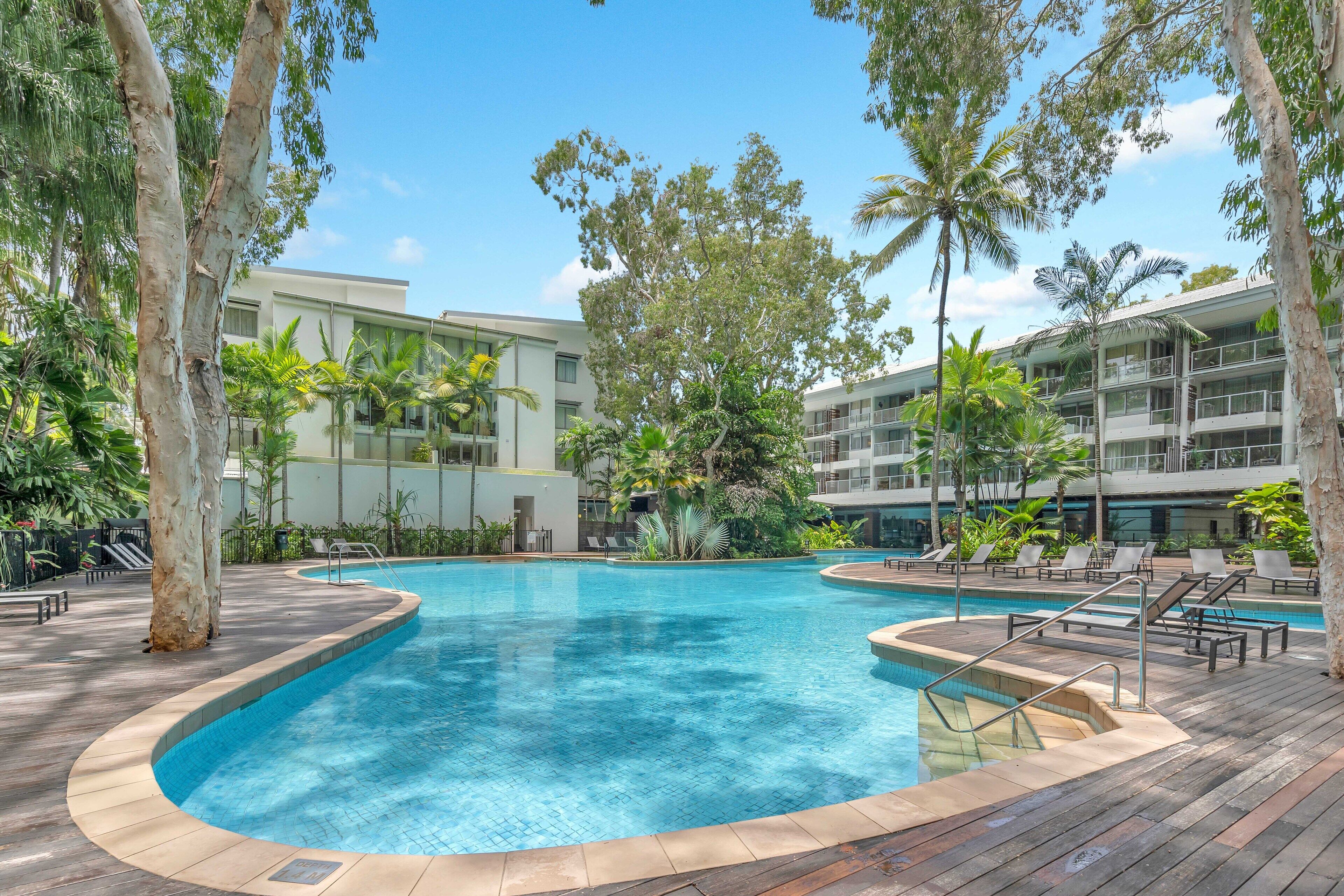A swimming pool surrounded by palm trees and lounge chairs, with a modern building in the background.