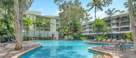 A swimming pool surrounded by palm trees and lounge chairs, with a modern building in the background.