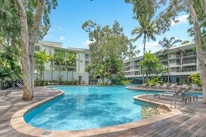 A swimming pool surrounded by palm trees and lounge chairs, with a modern building in the background.