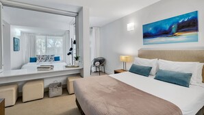 A modern hotel room with a large bed, a bedside table, a chair, a desk, a large mirror, and a painting on the wall. - Elysium The Drift Apartment (Palm Cove)