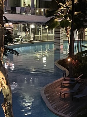 A swimming pool with a wooden deck and lounge chairs, surrounded by palm trees and a building in the background.