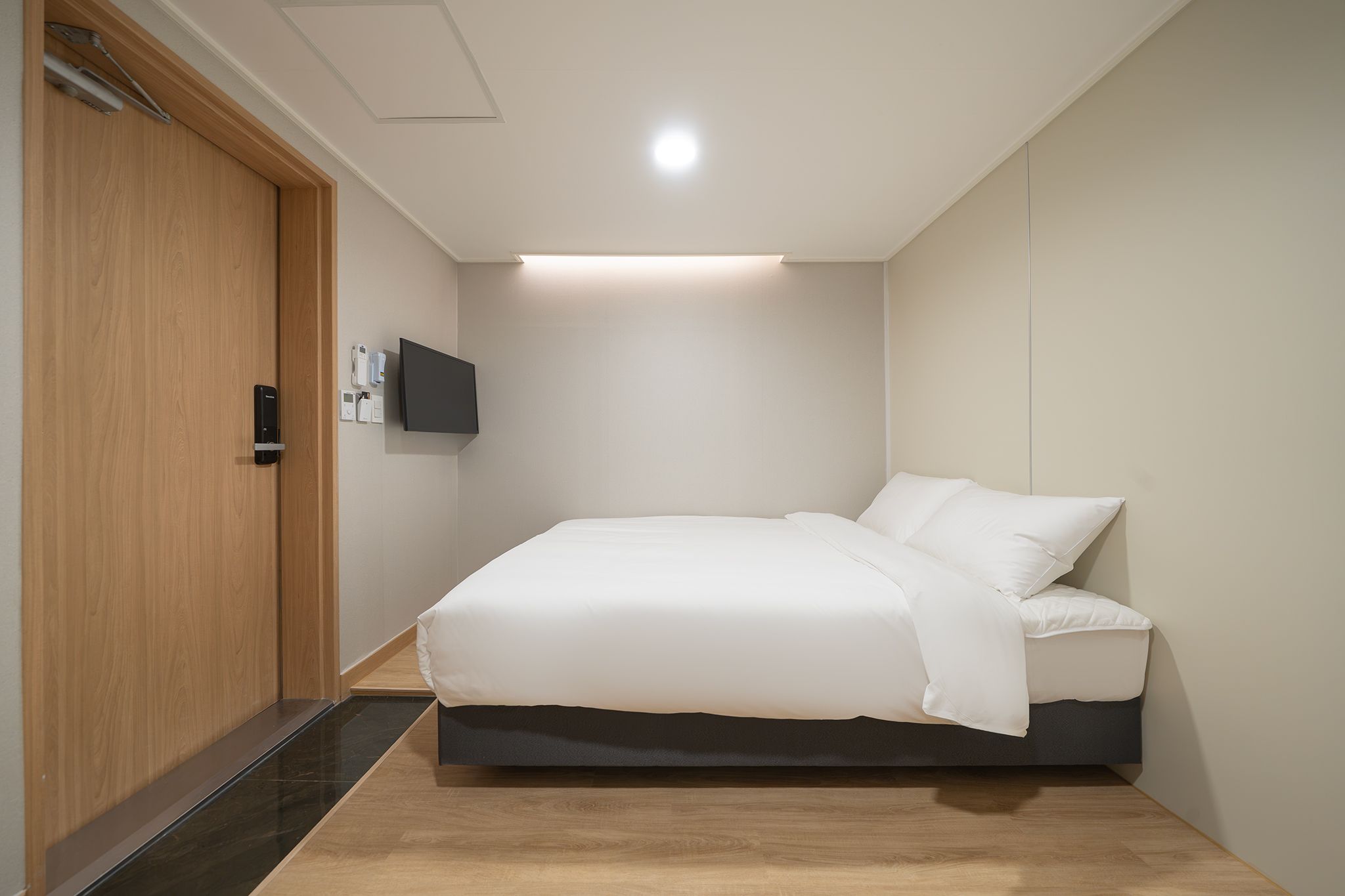 A bedroom with a bed, a wooden door, a wall-mounted TV, and a ceiling light.