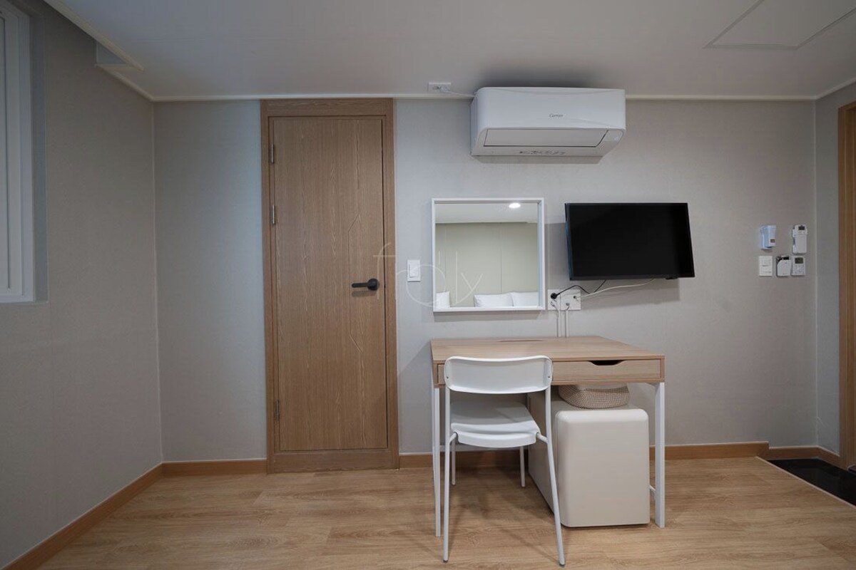 Una habitación con puerta de madera, un escritorio blanco, una silla, un televisor montado en la pared y un equipo de aire acondicionado.