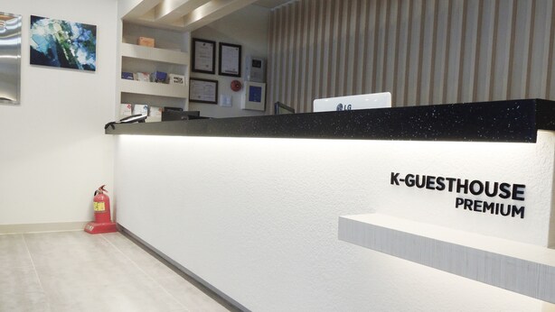 Una recepción con el rótulo "K-GUESTHOUSE PREMIUM", diseño moderno, un extintor y certificados enmarcados en la pared.