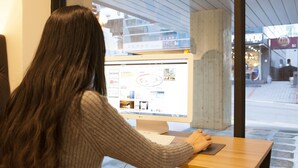 Una persona usando una computadora de escritorio en una mesa de madera en una cafetería con ventanas amplias.