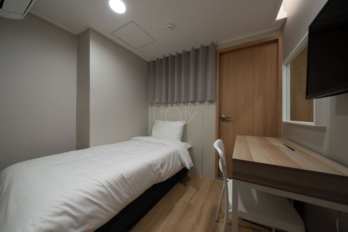 Una habitación pequeña con cama individual, un escritorio y una silla de madera, una ventana con cortinas y una puerta.