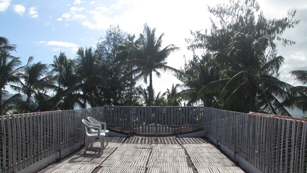 Una terraza de madera con una silla de plástico blanca, rodeada por una valla metálica, con vista a palmeras y una masa de agua.