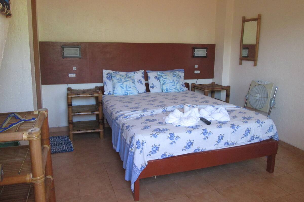 A bedroom with a wooden bed, two nightstands, a wooden chair, a small table, and a wall-mounted fan.