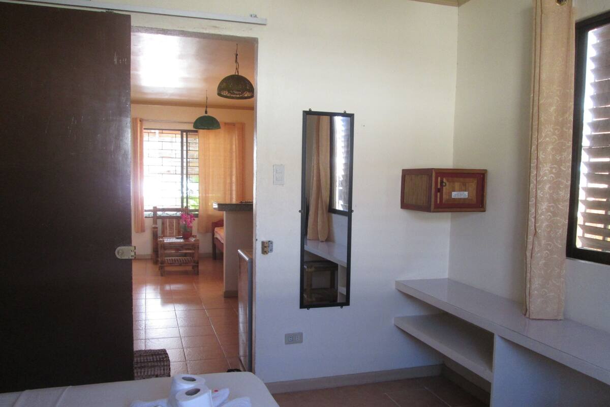A hotel room with a bed, a small table with toiletries, a mirror, and a view of the dining area.