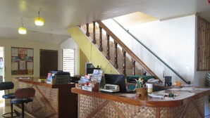 Zona de recepción con mostrador de madera, una computadora y un teléfono. Se observa una escalera al fondo.